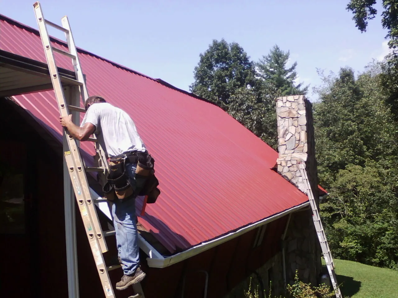 Roofer on a ladder beside a red metal roof and stone chimney — Soffit & Fascia Repair by Roofing Brothers in Neihart